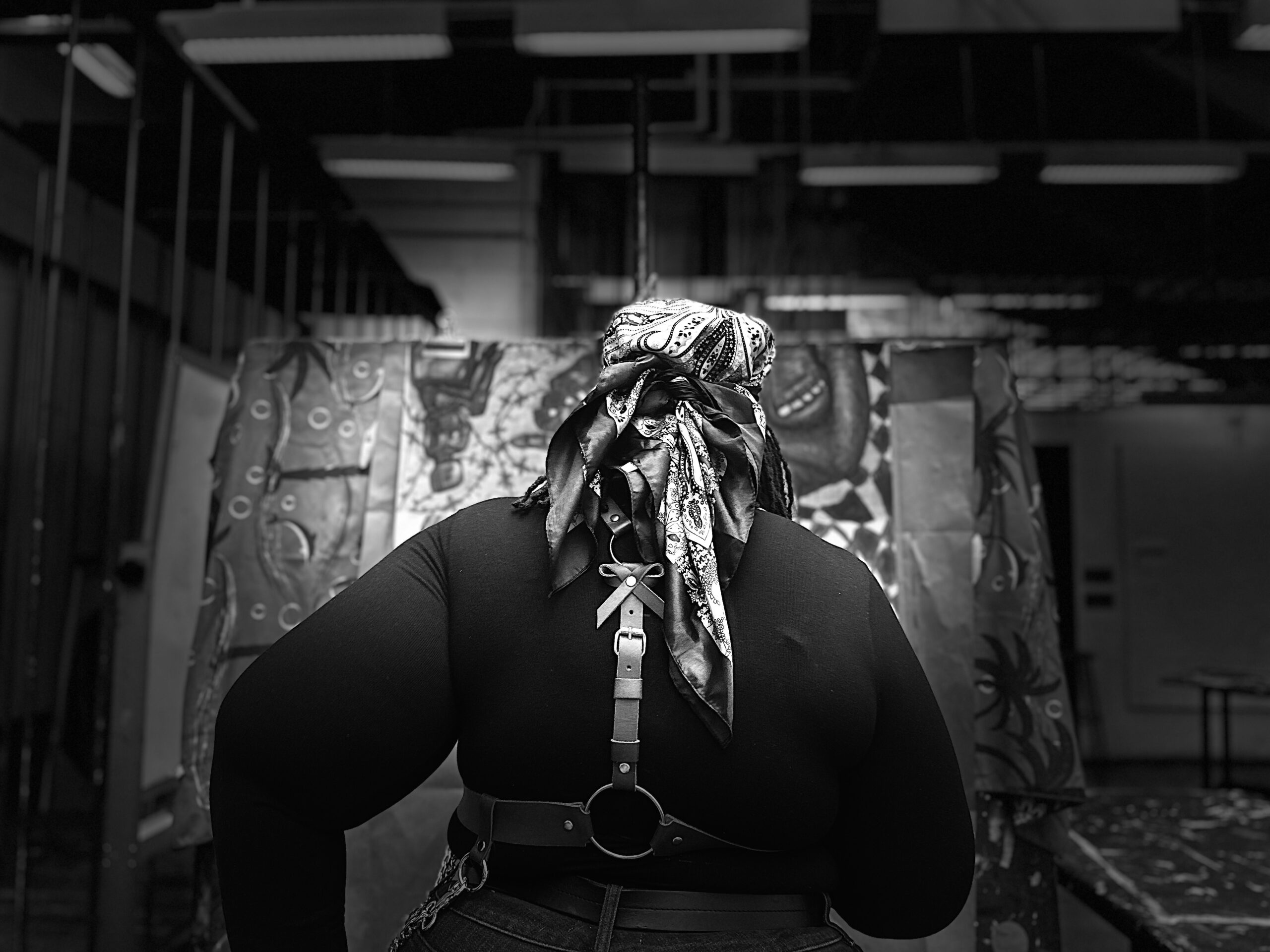 A person wearing a patterned headscarf and black outfit with a leather harness stands facing a large painting in an art studio. The image is in black and white, showing strong contrast between the figure and the creative background.